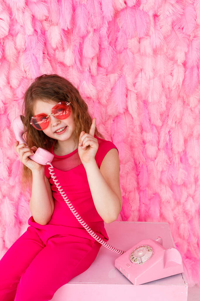 A little girl is sitting in stylish pink glasses on a pink background of their feathers. A beautiful baby girl is holding an old pink phone and tells something joyfully and holds a finger up - Photo, Image