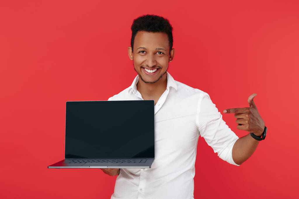 Mutlu Afrikalı Amerikalı genç adam dizüstü bilgisayarını tutuyor ve kırmızı arkaplanı işaret ediyor.. - Fotoğraf, Görsel