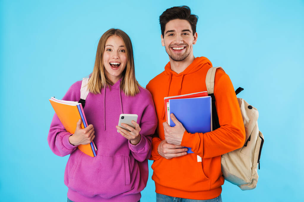Portrét veselého mladého páru studentů v batohech, nesoucích učebnice stojící nad modrým pozadím - Fotografie, Obrázek