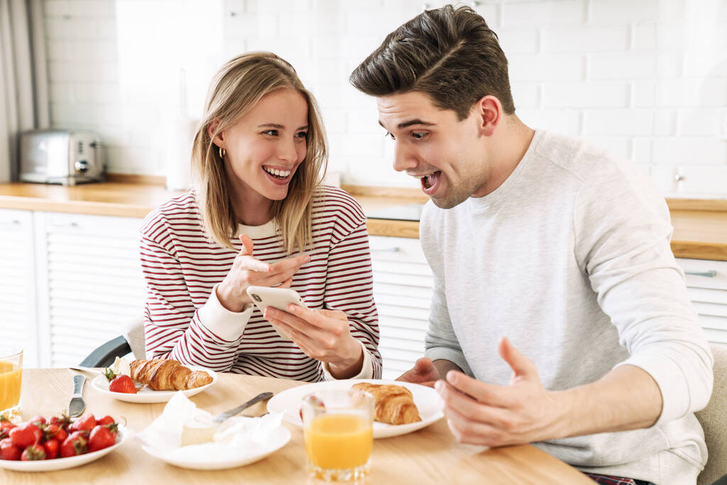 Портрет молодой счастливой пары с помощью смартфона во время завтрака в уютной кухне дома
 - Фото, изображение
