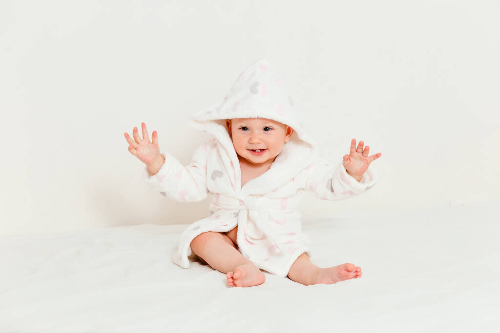 陽気な赤ちゃん女の子でAフード付きのローブでベッドの上に白い背景に座って - 写真・画像