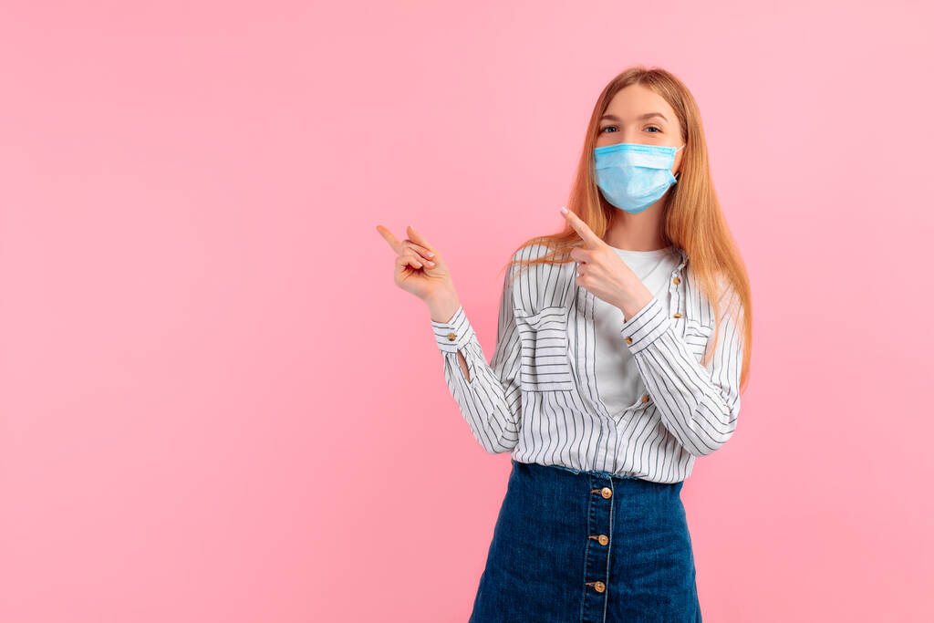 ελκυστική νεαρή γυναίκα σε μια ιατρική προστατευτική μάσκα στο πρόσωπό της, δείχνει το δάχτυλό της σε copyspace, σε ένα απομονωμένο ροζ φόντο - Φωτογραφία, εικόνα