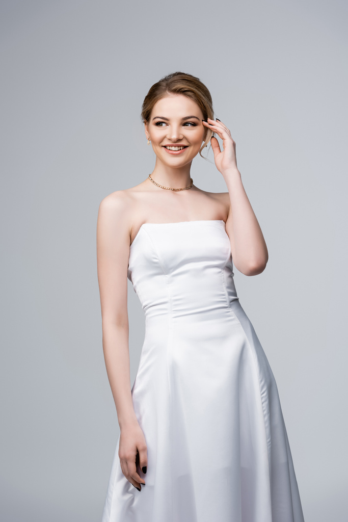 noiva feliz no vestido de casamento branco tocando o cabelo isolado no cinza
  - Foto, Imagem