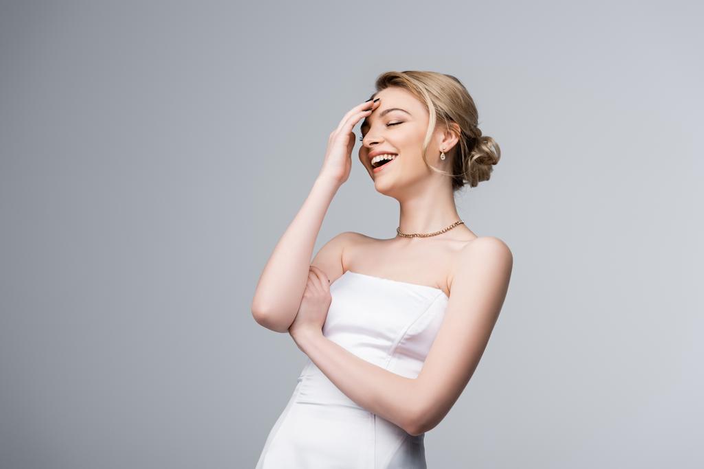 happy bride in white wedding dress laughing while touching forehead isolated on grey - Photo, Image