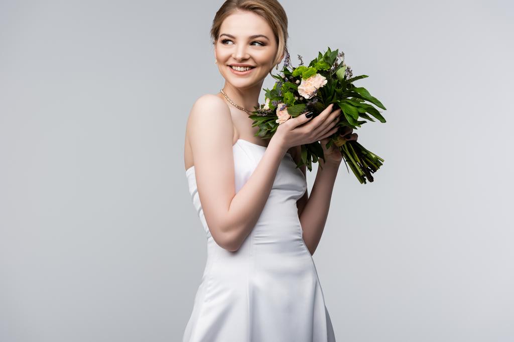 χαρούμενη νύφη με λευκό φόρεμα που κρατά τα λουλούδια του γάμου απομονωμένα σε γκρι  - Φωτογραφία, εικόνα