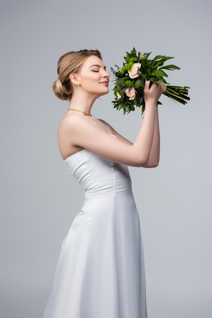 positive bride in white dress smelling wedding flowers isolated on grey  - Photo, Image