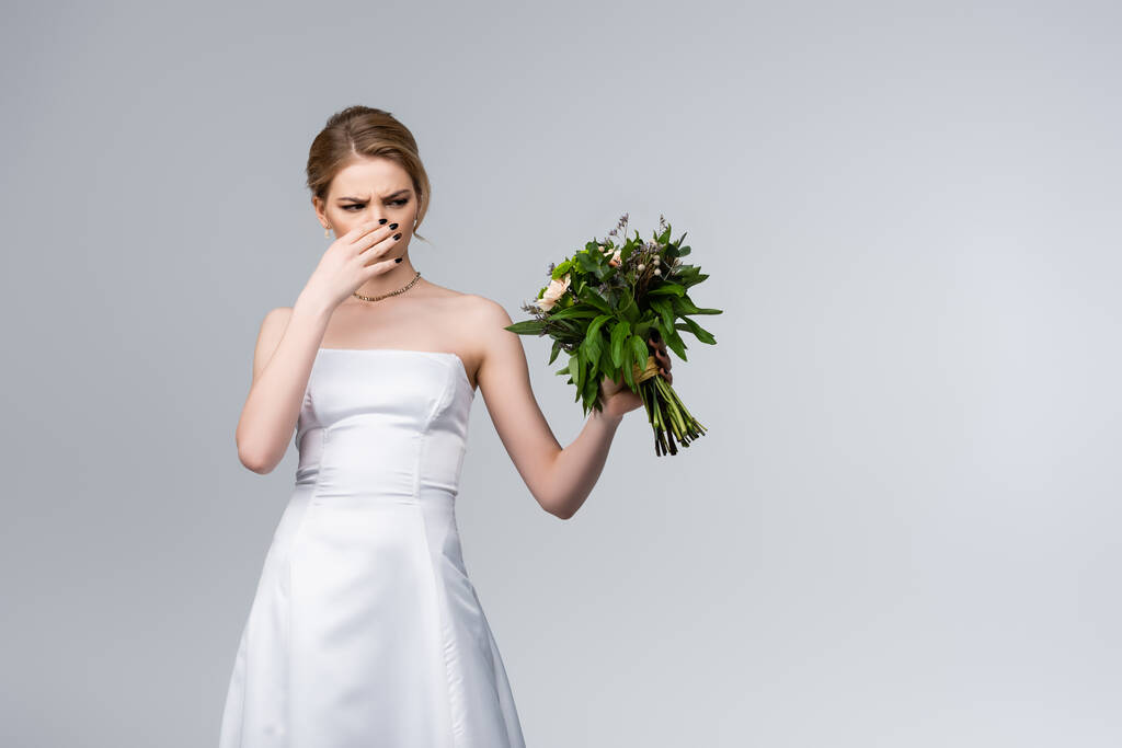 novia en vestido de novia blanco tocando la nariz mientras sostiene flores olorosas aisladas en gris
  - Foto, imagen