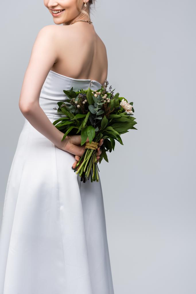 bijgesneden uitzicht op jonge en vrolijke vrouw in witte trouwjurk met boeket bloemen geïsoleerd op grijs  - Foto, afbeelding