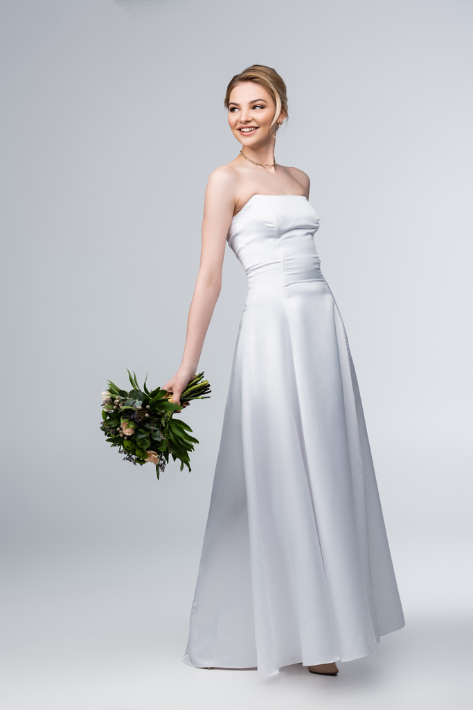 молодая и счастливая невеста в белом свадебном платье с букетом цветов на сером
  - Фото, изображение