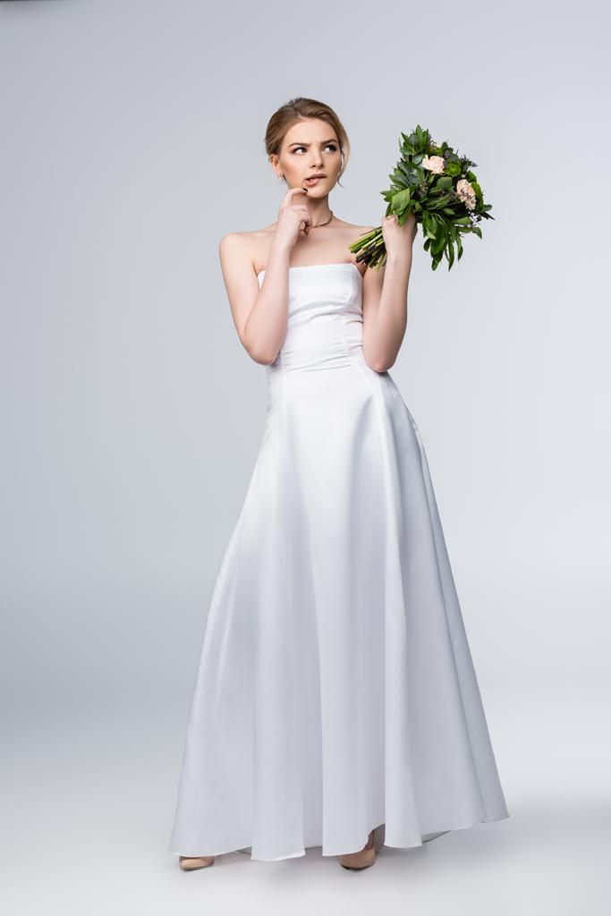 Ragazza pensierosa in abito da sposa bianco con mazzo di fiori e viso toccante sul grigio  - Foto, immagini