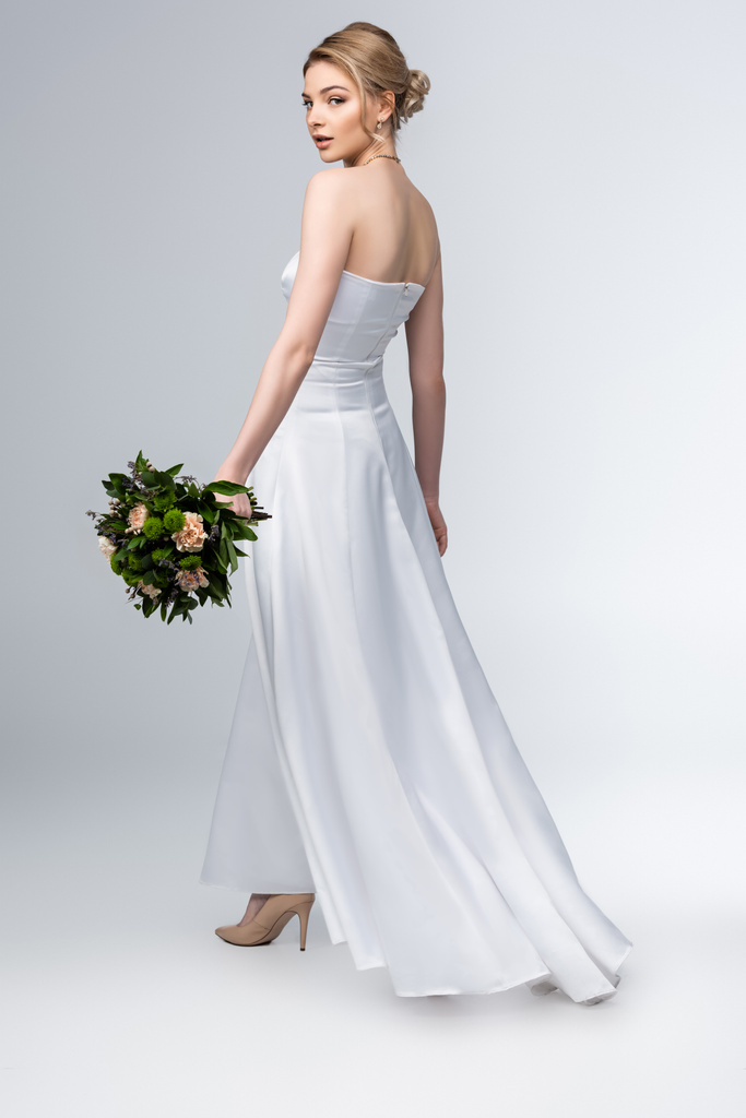 aantrekkelijke bruid in elegante trouwjurk met boeket bloemen op grijs - Foto, afbeelding