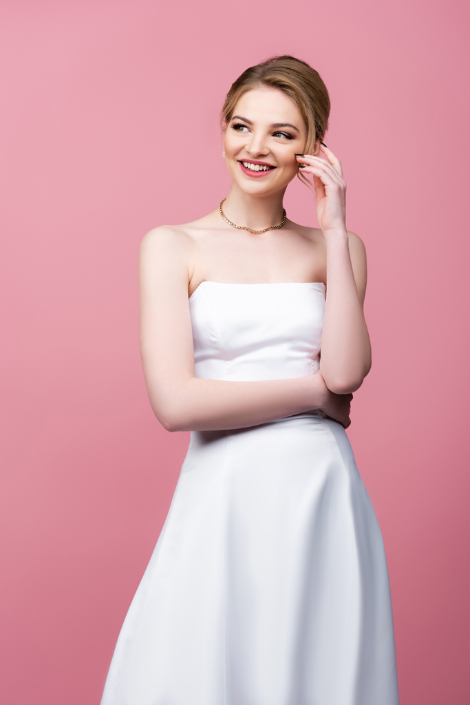 szczęśliwa panna młoda w białej sukni ślubnej dotykając twarzy odizolowany na różowy  - Zdjęcie, obraz