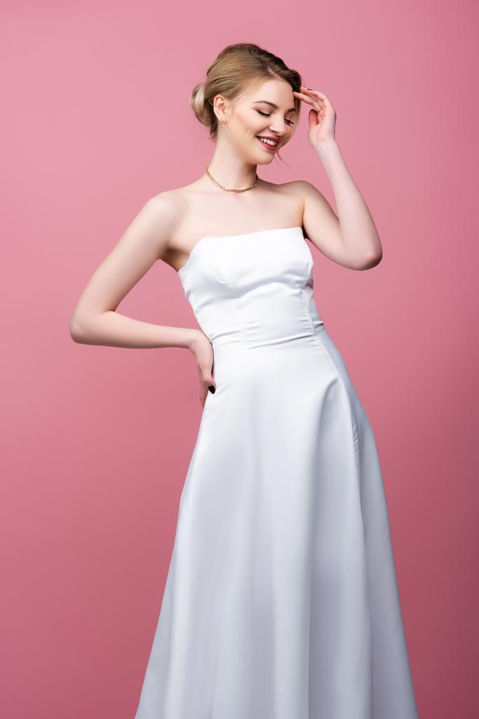 glückliche junge Braut im weißen Hochzeitskleid stehend mit der Hand an der Hüfte isoliert auf rosa  - Foto, Bild