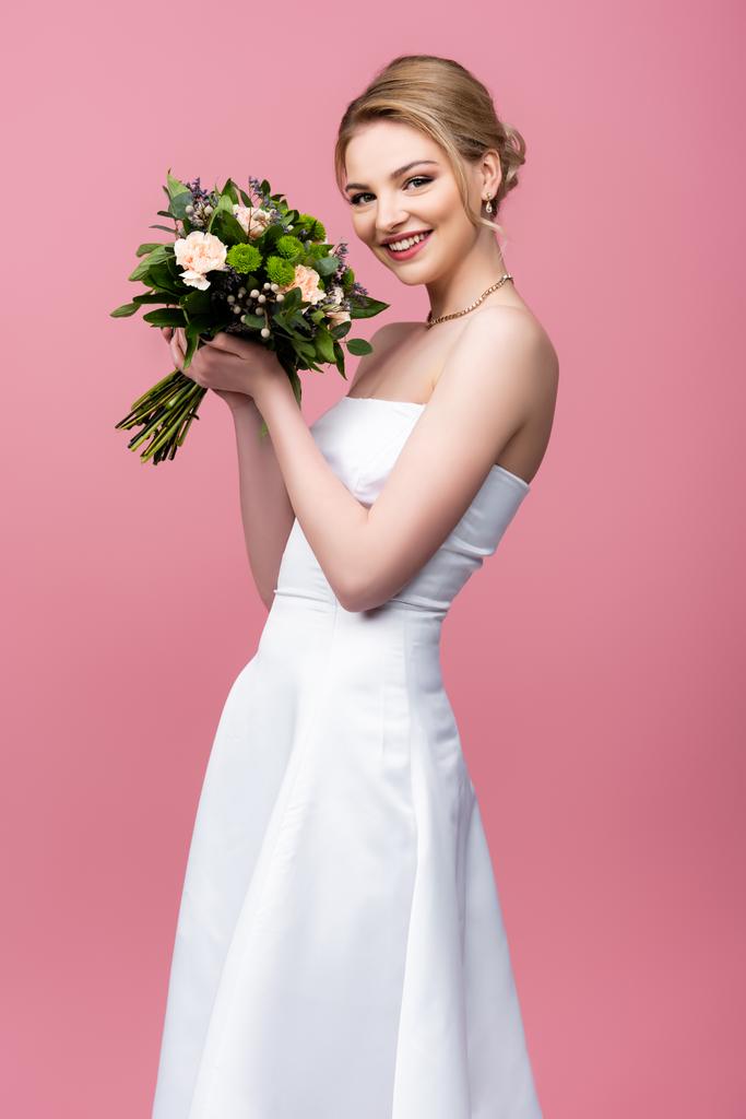 χαρούμενη νύφη σε λευκό νυφικό κρατώντας λουλούδια και κοιτάζοντας κάμερα απομονωμένη σε ροζ  - Φωτογραφία, εικόνα