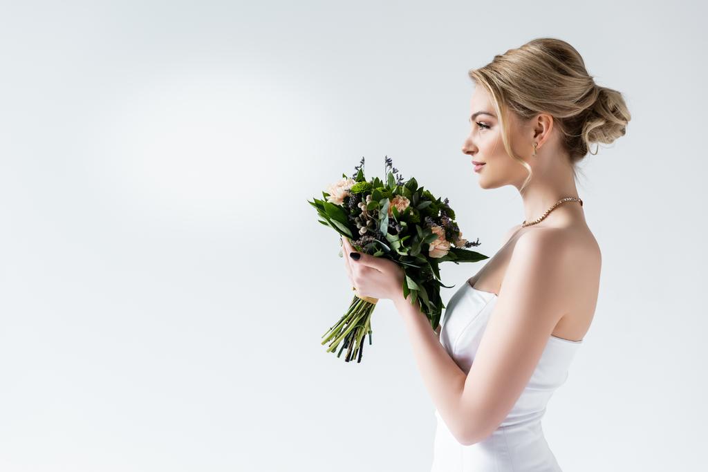 πλαϊνή όψη της ελκυστικής νύφης κρατώντας λουλούδια του γάμου σε λευκό  - Φωτογραφία, εικόνα