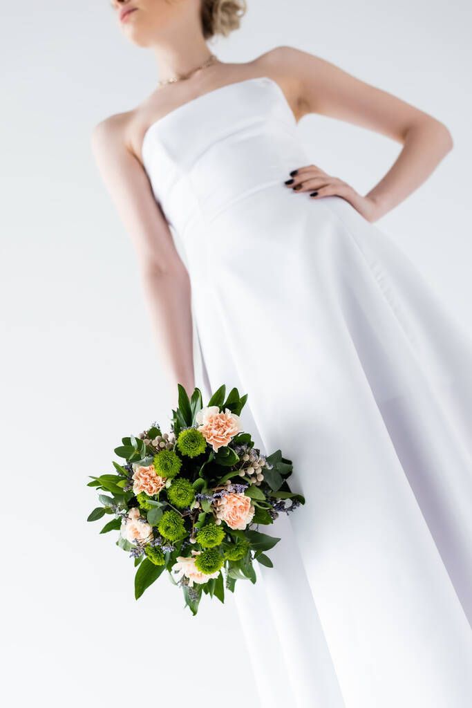 przycięty widok eleganckiej panny młodej w sukni ślubnej gospodarstwa kwiaty izolowane na białym  - Zdjęcie, obraz