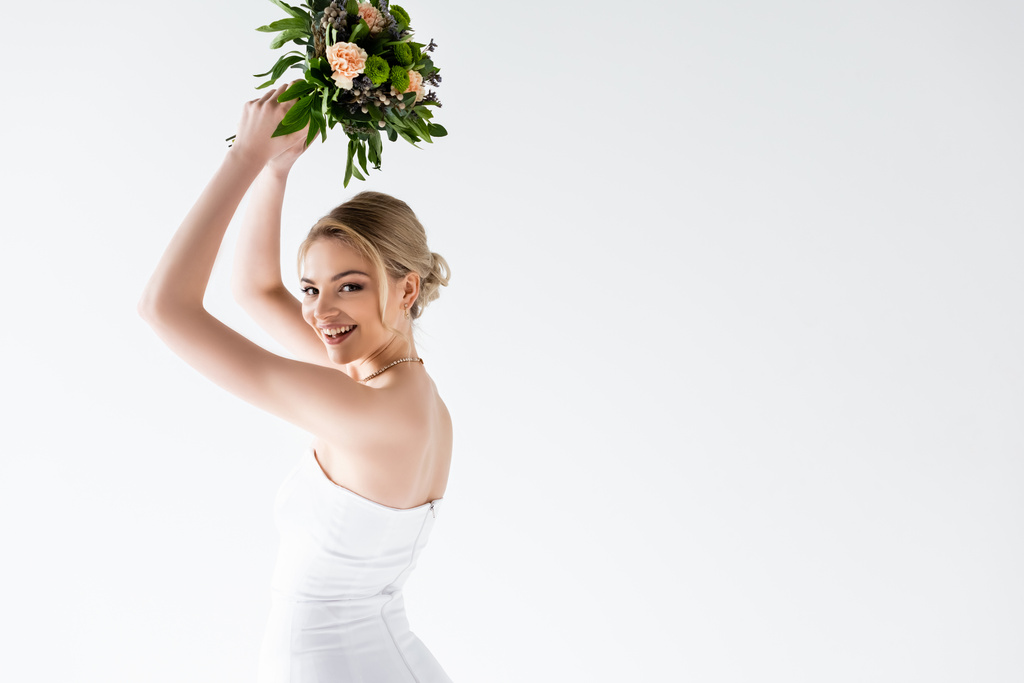 junge und fröhliche Braut im eleganten Brautkleid mit Blumen über dem Kopf isoliert auf weiß  - Foto, Bild