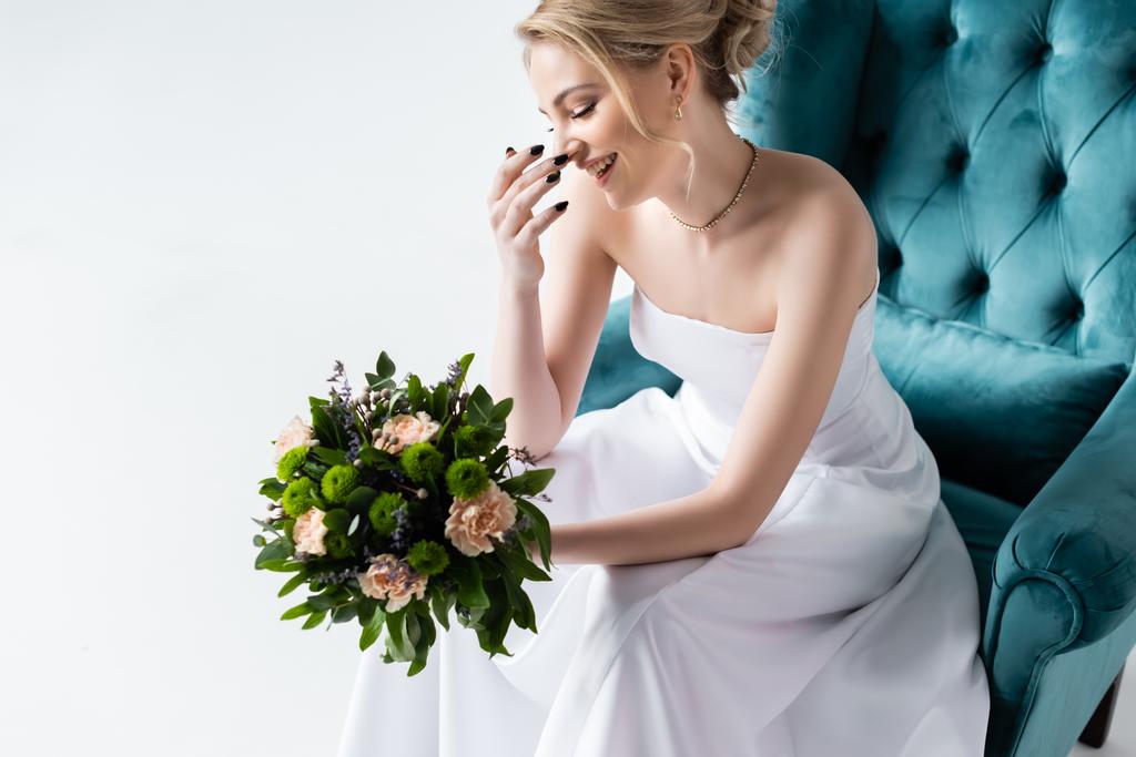 usmívající se nevěsta v elegantních svatebních šatech drží květiny, zatímco sedí v křesle a dotýká tvář izolované na bílém - Fotografie, Obrázek