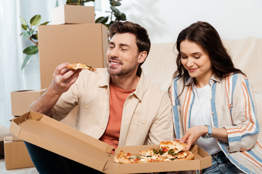 Χαμογελώντας άνθρωπος κρατώντας κομμάτι πίτσα κοντά στην όμορφη φίλη και κουτιά από χαρτόνι στο σπίτι - Φωτογραφία, εικόνα