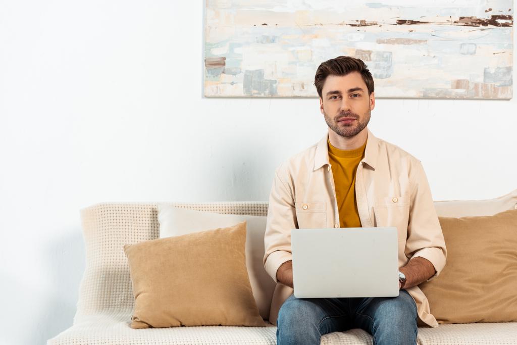 Handsome freelancer looking at camera while holding laptop on couch  - Photo, Image
