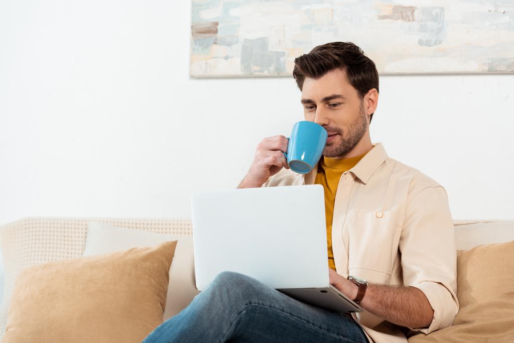 Guapo freelancer beber café mientras usa el ordenador portátil en la sala de estar
 - Foto, Imagen