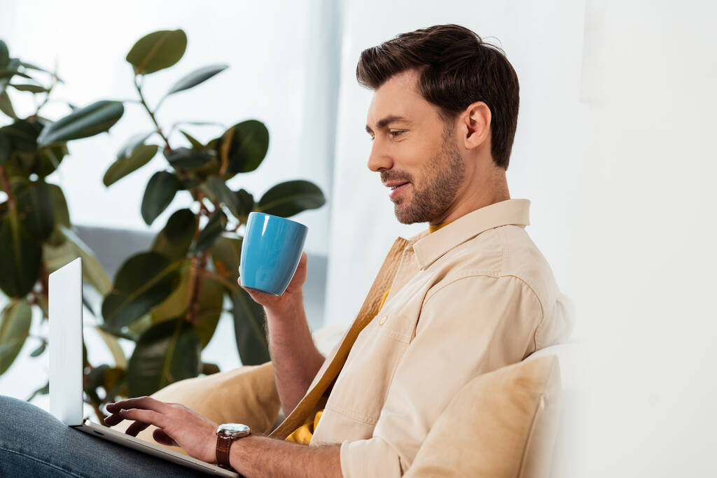 Επιλεκτική εστίαση του όμορφου άνδρα κρατώντας φλιτζάνι καφέ και χρησιμοποιώντας φορητό υπολογιστή στο σπίτι  - Φωτογραφία, εικόνα