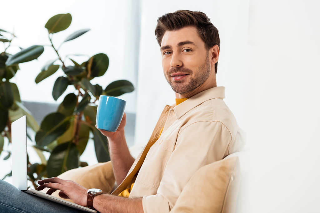 Side view of handsome freelancer holding cup of coffee and using laptop in living room - Photo, Image