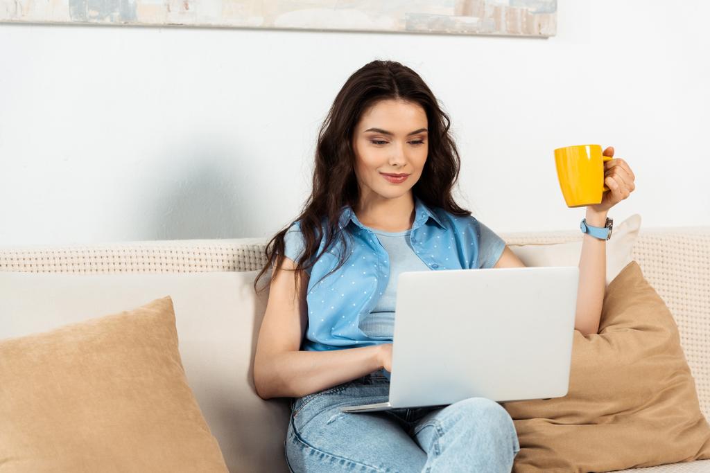 Attractive freelancer holding coffee cup and using laptop in living room - Photo, Image