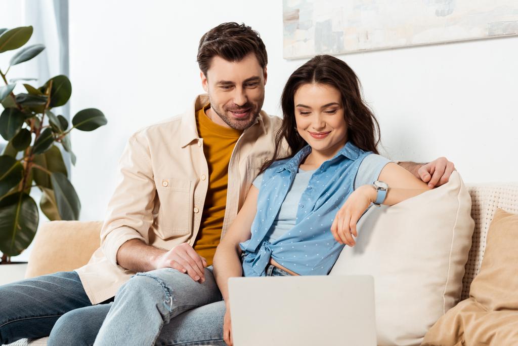 Selective focus of handsome man sitting near smiling girlfriend and laptop on couch  - Photo, Image