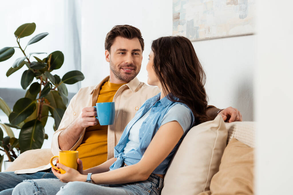 Szelektív fókusz mosolygós férfi iszik kávét közelében barátnője otthon  - Fotó, kép