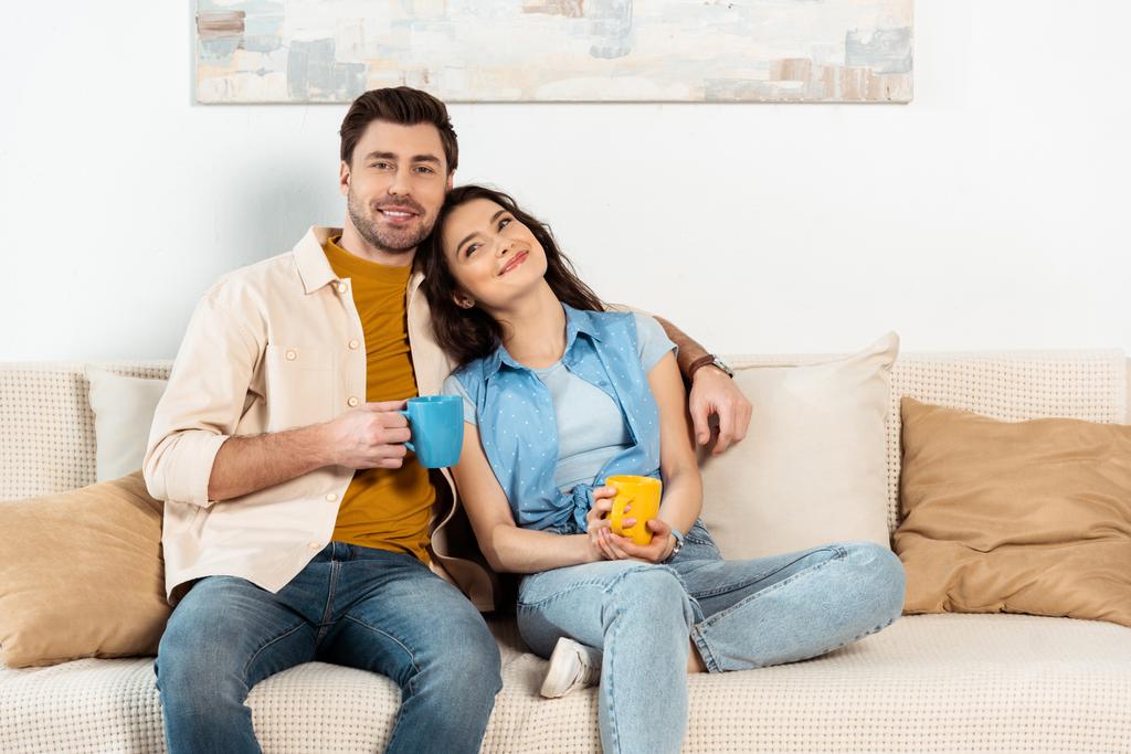 Schöner Mann lächelt in die Kamera, während er Freundin umarmt und eine Tasse Kaffee auf dem Sofa hält  - Foto, Bild