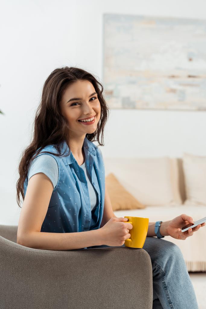 Gyönyörű nő mosolyog a kamera használata közben okostelefon és a kezében csésze kávét karosszékben  - Fotó, kép