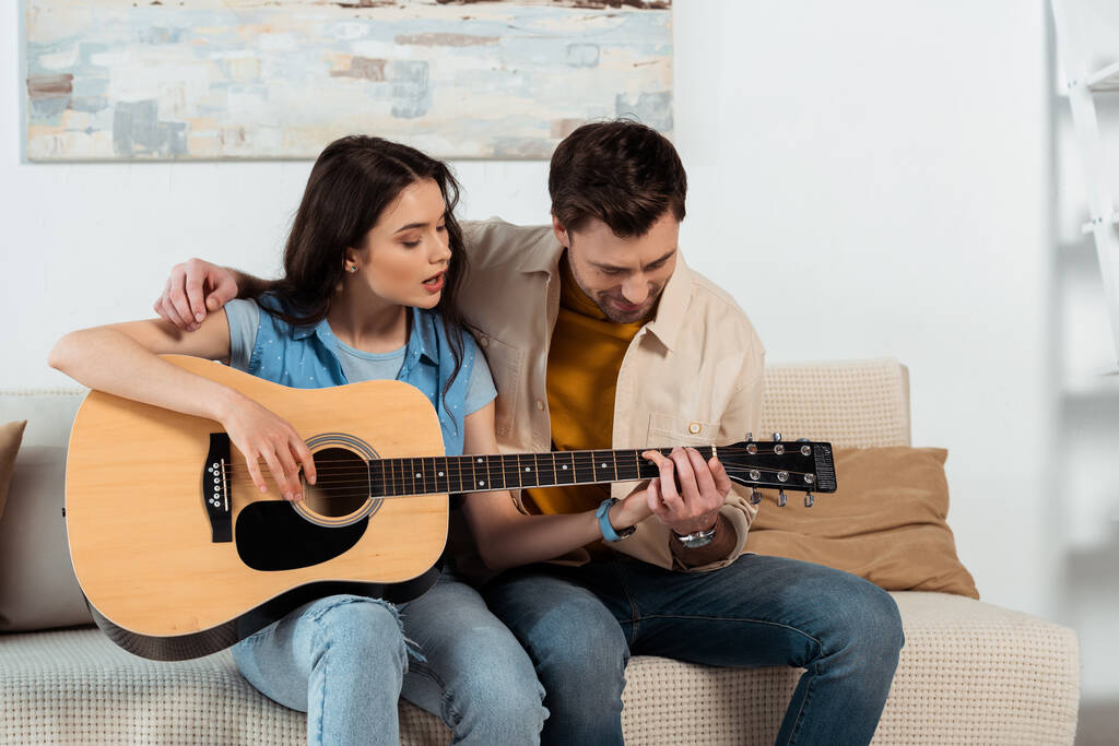 Мужчина учит девушку играть на акустической гитаре в гостиной
 - Фото, изображение