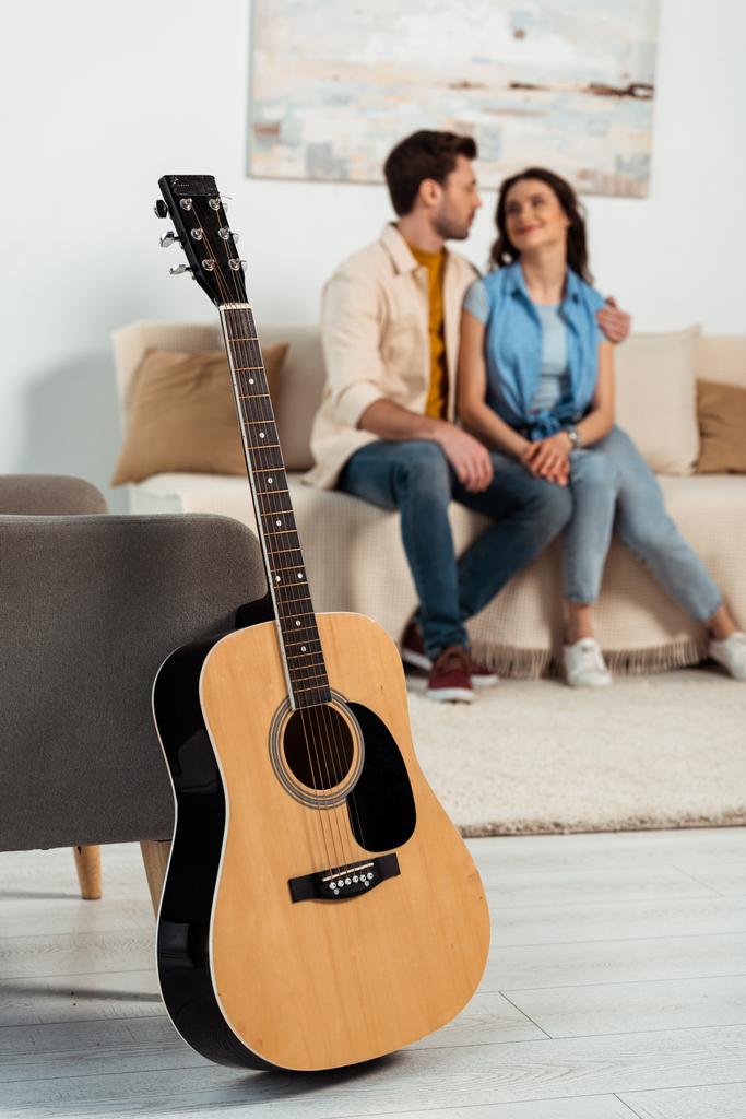Selektywne skupienie gitary akustycznej na podłodze w pobliżu fotela i mężczyzna obejmujący dziewczynę w domu  - Zdjęcie, obraz