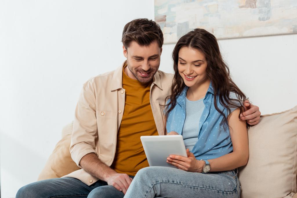Komea mies syleilee hymyilevä tyttöystävä, kun käytät digitaalista tablettia kotona  - Valokuva, kuva