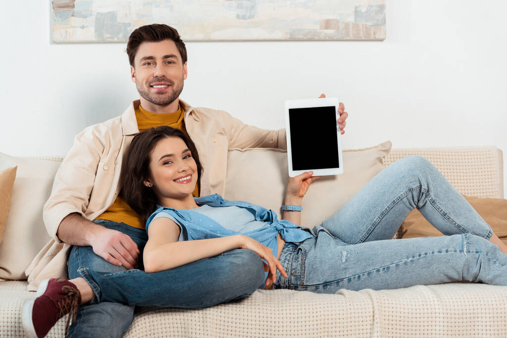 Νεαρό ζευγάρι χαμογελά στην κάμερα, ενώ δείχνει ψηφιακή ταμπλέτα με λευκή οθόνη στο σαλόνι - Φωτογραφία, εικόνα