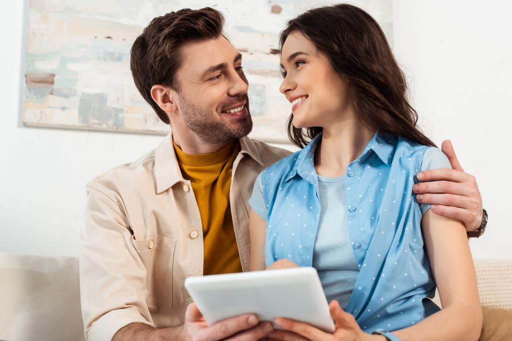 Selective focus of man hugging smiling woman with digital tablet at home  - Photo, Image