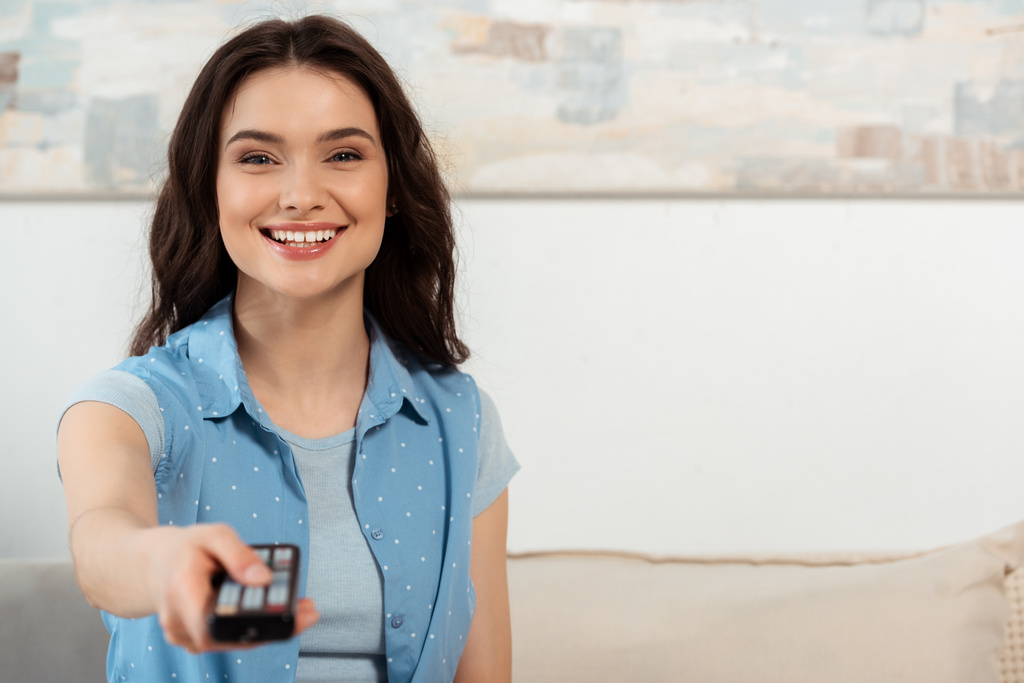 Enfoque selectivo de la hermosa mujer sonriendo a la cámara mientras sostiene el mando a distancia en casa
  - Foto, Imagen