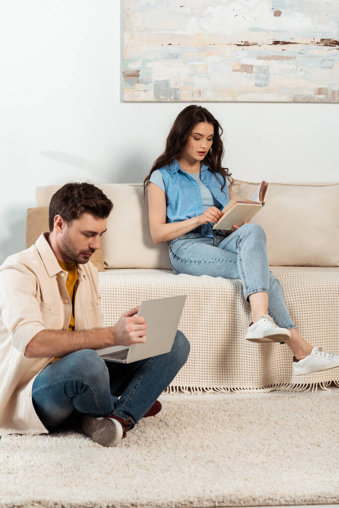 Muž pomocí notebooku poblíž ženy čtení knihy v obývacím pokoji - Fotografie, Obrázek
