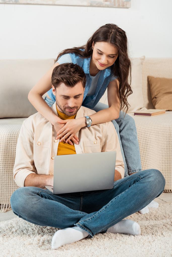 Smiling woman hugging boyfriend using laptop at home  - Photo, Image