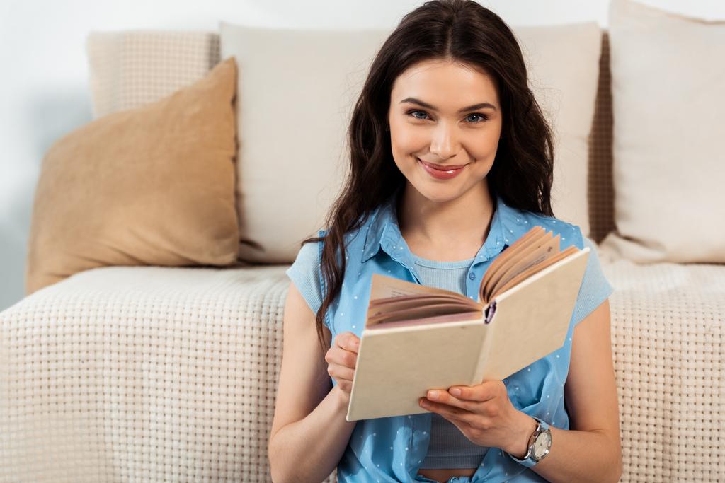 Молода жінка посміхається на камеру, читаючи книгу у вітальні
 - Фото, зображення