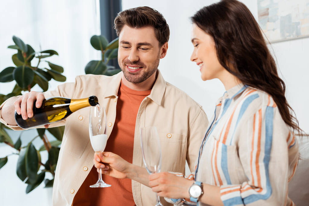 Selective focus of smiling man pouring champagne in glass near beautiful girlfriend at home  - 写真・画像