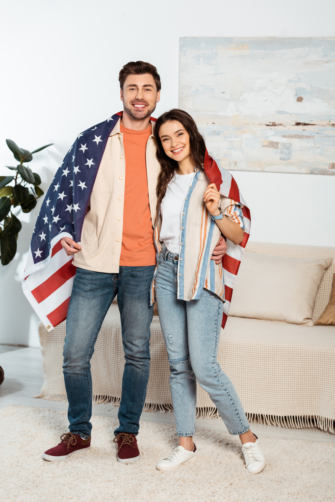 Junges Paar hüllt sich in amerikanische Flagge und lächelt in Wohnzimmer in die Kamera - Foto, Bild