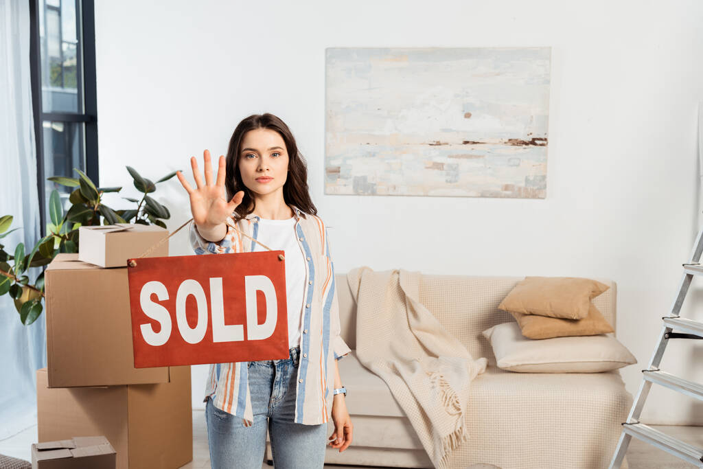 Mladá žena drží jmenovku s prodaným nápisem v blízkosti lepenkových krabic v obývacím pokoji - Fotografie, Obrázek
