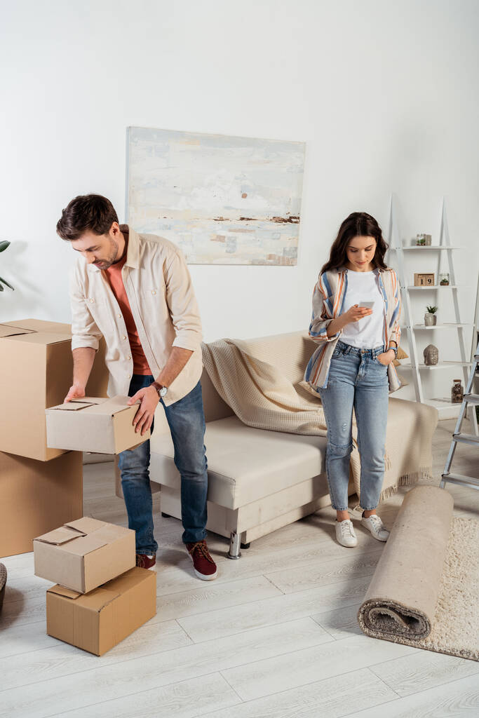 Man met kartonnen doos terwijl vriendin met behulp van smartphone in de woonkamer tijdens het verplaatsen  - Foto, afbeelding