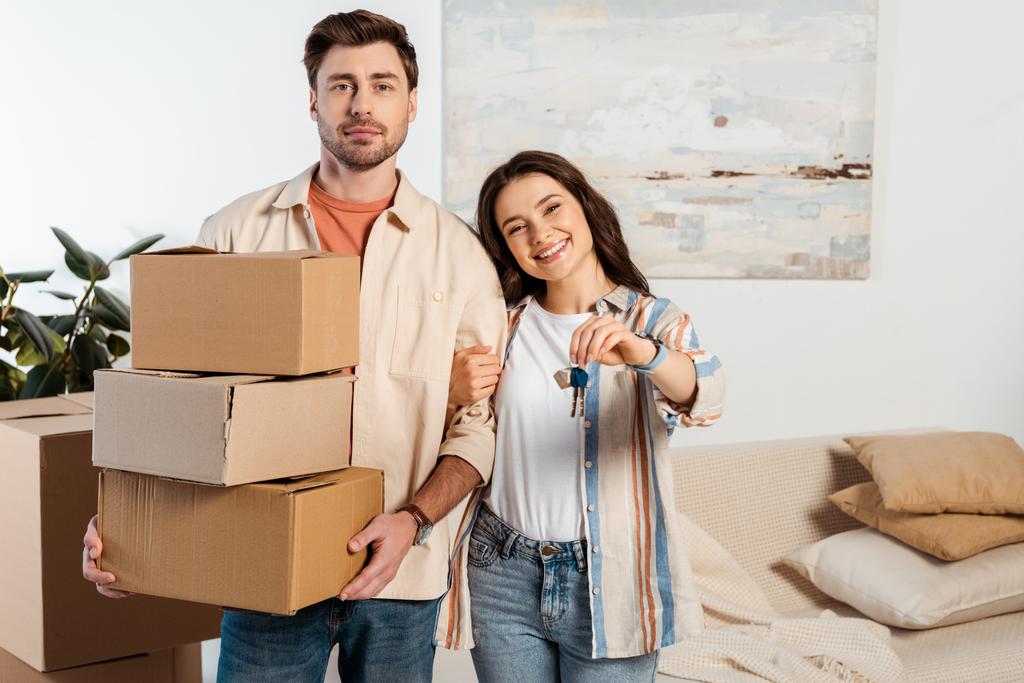 Lächelnde Frau hält Schlüssel für neues Haus neben Freund mit Kartons zu Hause  - Foto, Bild