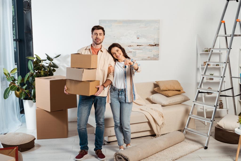Positive woman holding keys of new house near boyfriend with cardboard boxes in living room - Photo, Image