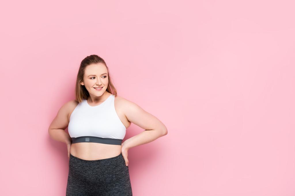 cheerful overweight girl in sportswear holding hands on hips and looking away on pink - Photo, Image