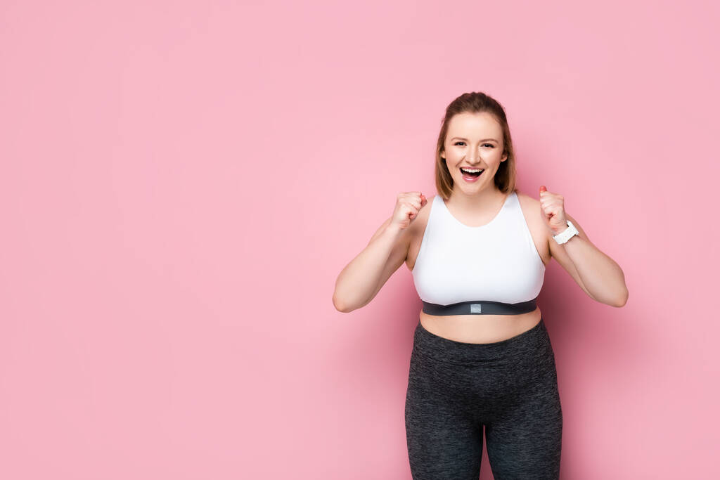 Aufgeregtes übergewichtiges Mädchen in Sportbekleidung zeigt Siegergeste auf rosa - Foto, Bild
