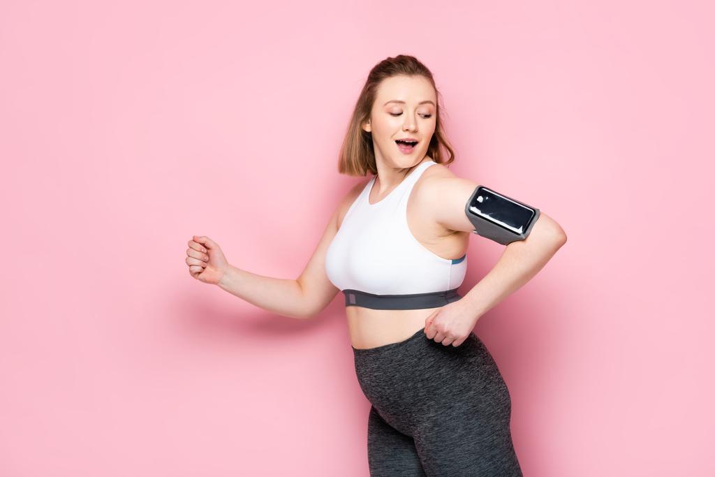 sorridente ragazza in sovrappeso che corre mentre guarda lo smartphone in fascia su rosa - Foto, immagini